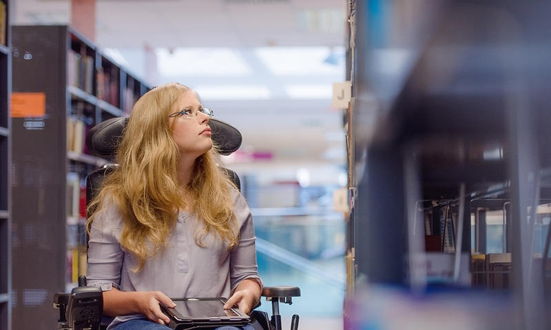 student in a wheelchair with a tablet computer looks at library shelves photo by istock